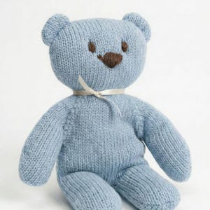 May include: A light blue knitted teddy bear featuring a smooth, textured yarn surface. The bear has two small dark brown embroidered eyes and a triangular dark brown embroidered nose. A thin, cream-colored ribbon is tied in a bow around its neck.