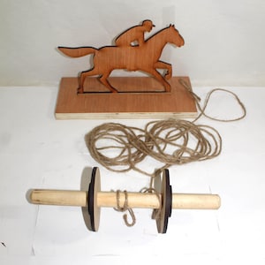 May include: A wooden toy depicting a horse and jockey, cut from wood and mounted on a wooden base. A spool-shaped wooden toy with dark brown circular ends and a light brown handle is connected to the horse by twine. A children's toy.