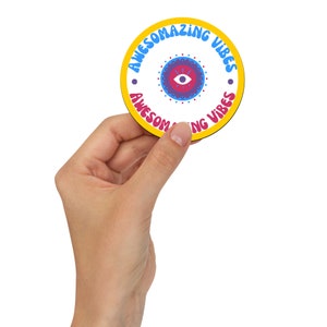 Könnte beinhalten: Ein runder Untersetzer mit gelbem Rand und den Worten "AWESOMAZING VIBES" in Blau und Pink. In der Mitte befindet sich ein Design mit einem Augensymbol in Rosa-, Blau- und Rottönen. Der Untersetzer wird von einer Hand gehalten.