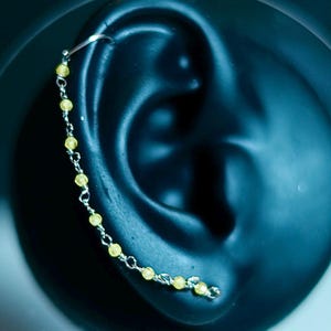 May include: A silver ear cuff with a delicate chain of small, yellow beads. The ear cuff curves along the ear, with the chain of beads dangling down. The background is a dark, blurred shape, possibly an ear.