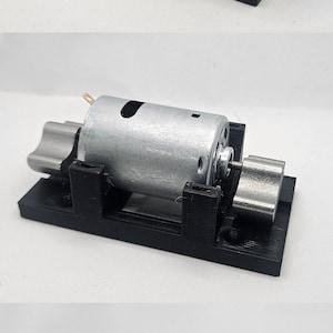 以下が含まれることがあります： 銀色の電動モーターが取り付けられた3Dプリントのモーターマウント。モーターマウントは黒で、モーターの両端に黒いプラスチックリングが2つあります。