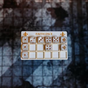 May include: A white dice holder with the words "FAITH DICE" in gold. It holds six pearlised dice with gold pips and a fleur-de-lis. The dice are arranged in a grid pattern. The background is a patterned surface.