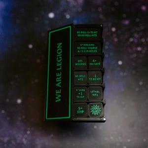 May include: A black rectangular game tracker with green text and icons. The left side features the text WE ARE LEGION. The right side contains a grid of small square tiles with various game rules and stats printed in green, including re-roll hits.