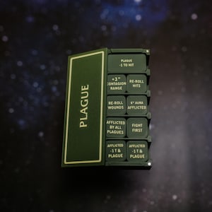 May include: A dark olive green rectangular game token holder with a hinged lid labeled PLAGUE. Inside are ten square tokens featuring white text including PLAGUE -1 TO HIT, +3" CONTAGION RANGE, RE-ROLL HITS, RE-ROLL WOUNDS, 6" AURA AFFLICTED, FIGHT FIRST, and AFFLICTED -1T & PLAGUE.