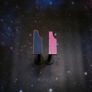 May include: Two abstract, geometric sculptures. One is navy blue with a pink outline, the other is pink with a pink outline. Both are mounted on dark gray stands against a dark, speckled background.