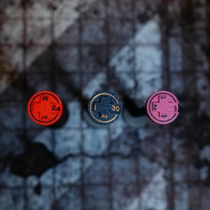 May include: Three circular game tokens with numbers and symbols. The tokens are red, blue, and pink, each with a different number. The red token has the number 24, the blue token has the number 30, and the pink token has the number 5.