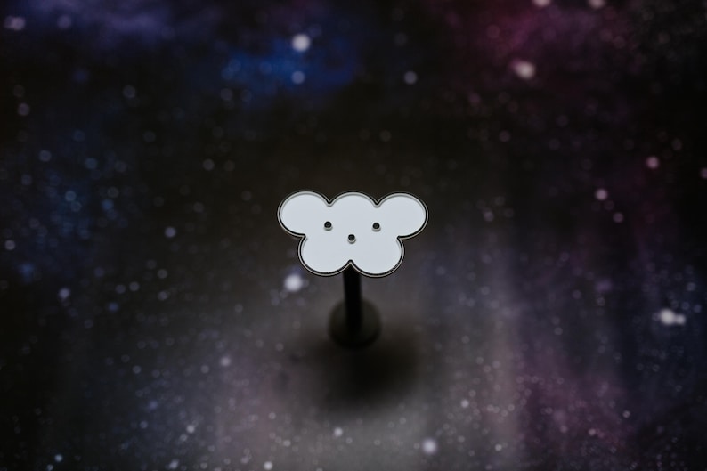 May include: A white cloud-shaped enamel pin with a black outline and two small black dots for eyes. The pin is mounted on a dark metal post, set against a starry galaxy background.