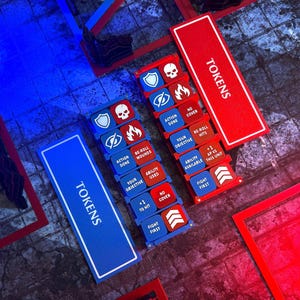 May include: Two rectangular game token organisers in red and blue, each labelled TOKENS. The organisers hold square tiles with white icons and text including shield, skull, eye, flame, and chevron symbols, plus labels like ACTION DONE and FIGHT FIRST.