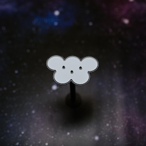 May include: A white cloud-shaped enamel pin with a black outline and three small black dots for eyes. The pin is mounted on a black post, set against a dark background with a galaxy-like pattern.