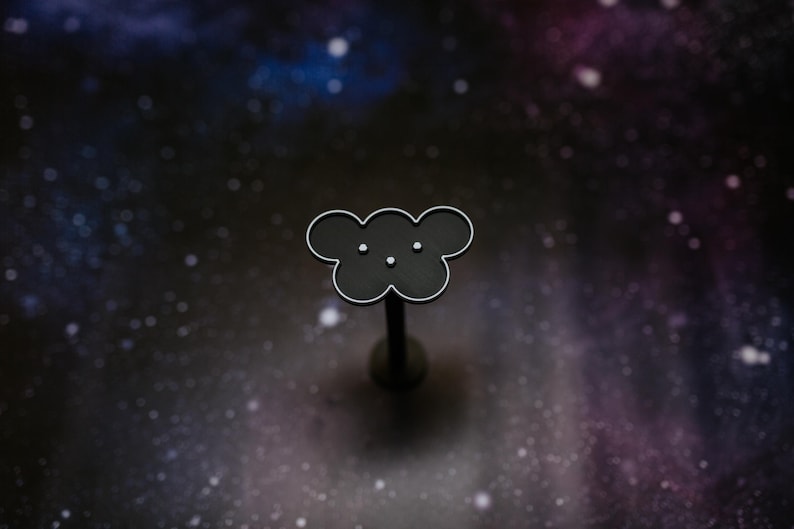 May include: A black cloud-shaped pin with a silver outline and three small white dots, set against a dark, starry background. The pin is mounted on a thin, dark post. The design is simple and elegant.