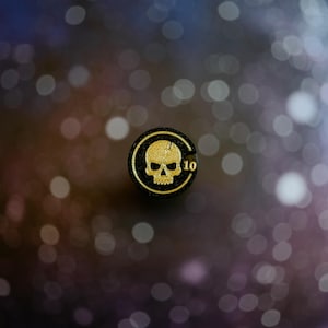 May include: A circular black token featuring a gold-colored skull graphic centered within a thin gold border. The number 10 is printed in gold on the right side of the skull. The token has a textured, glittery finish across its surface.