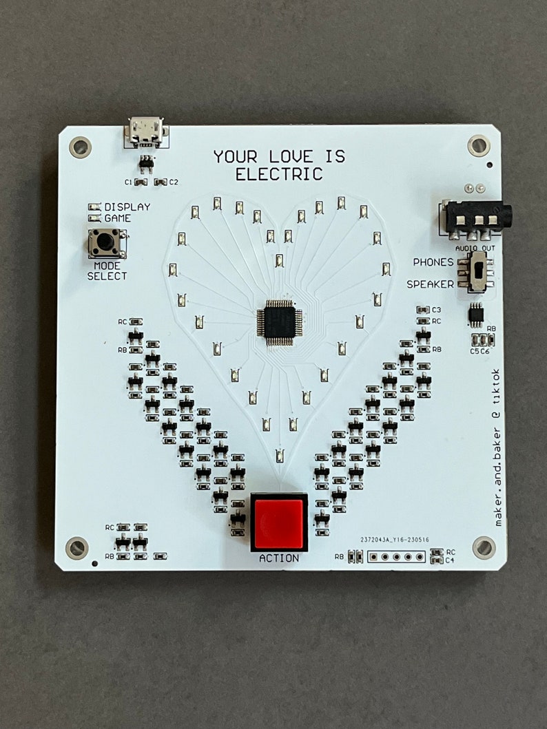 K&ouml;nnte beinhalten: Eine wei&szlig;e Platine mit einem herzf&ouml;rmigen Muster aus LEDs. Die Platine hat einen roten Knopf mit der Aufschrift "ACTION", einen Knopf mit der Aufschrift "MODE SELECT" und einen Knopf mit der Aufschrift "DISPLAY GAME". Die Platine hat au&szlig;erdem einen Lautsprecher, eine Kopfh&ouml;rerbuchse und einen USB-Anschluss. Der Text "YOUR LOVE IS ELECTRIC" ist auf der Platine aufgedruckt.