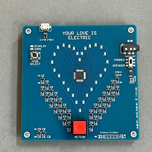 K&ouml;nnte beinhalten: Eine blaue Platine mit einem herzf&ouml;rmigen LED-Display, einem roten Knopf mit der Aufschrift "ACTION" und dem Text "YOUR LOVE IS ELECTRIC".