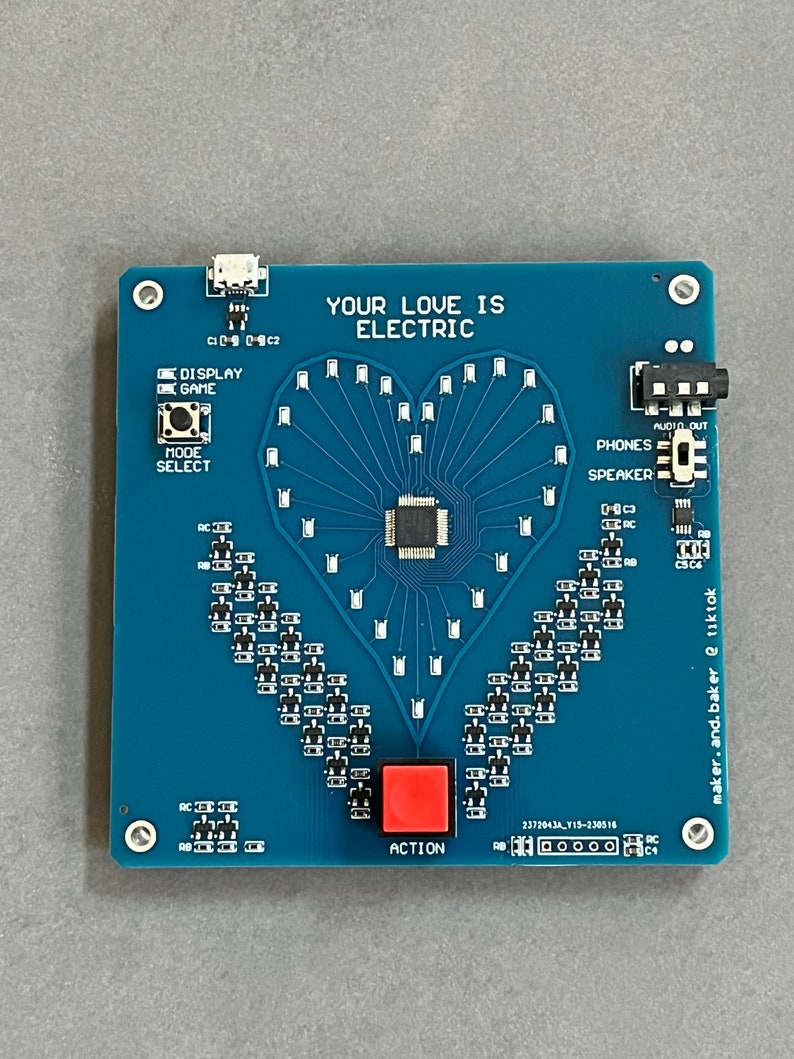 K&ouml;nnte beinhalten: Eine blaue Platine mit einem herzf&ouml;rmigen LED-Display, einem roten Knopf mit der Aufschrift "ACTION" und dem Text "YOUR LOVE IS ELECTRIC".