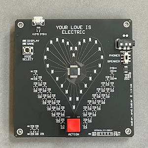 K&ouml;nnte beinhalten: Eine schwarze Platine mit einem herzf&ouml;rmigen Design aus LEDs. Die Platine hat einen roten Knopf mit der Aufschrift "ACTION", einen kleineren Knopf mit der Aufschrift "MODE SELECT" und einen Text, der "YOUR LOVE IS ELECTRIC" lautet.