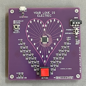 K&ouml;nnte beinhalten: Eine violette Leiterplatte mit einem herzf&ouml;rmigen Design aus LEDs. Die Platine hat einen roten Knopf mit der Aufschrift "ACTION", einen Lautsprecher und den Text "YOUR LOVE IS ELECTRIC".
