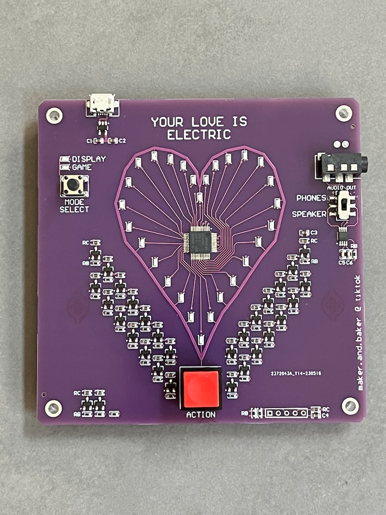 K&ouml;nnte beinhalten: Eine violette Leiterplatte mit einem herzf&ouml;rmigen Design aus LEDs. Die Platine hat einen roten Knopf mit der Aufschrift "ACTION", einen Lautsprecher und den Text "YOUR LOVE IS ELECTRIC".