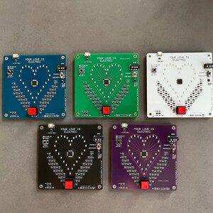 K&ouml;nnte beinhalten: F&uuml;nf Platinen mit dem aufgedruckten Text "Your Love Is Electric". Die Platinen sind in verschiedenen Farben: blau, gr&uuml;n, wei&szlig;, schwarz und lila. Jede Platine hat ein herzf&ouml;rmiges Muster aus LEDs.