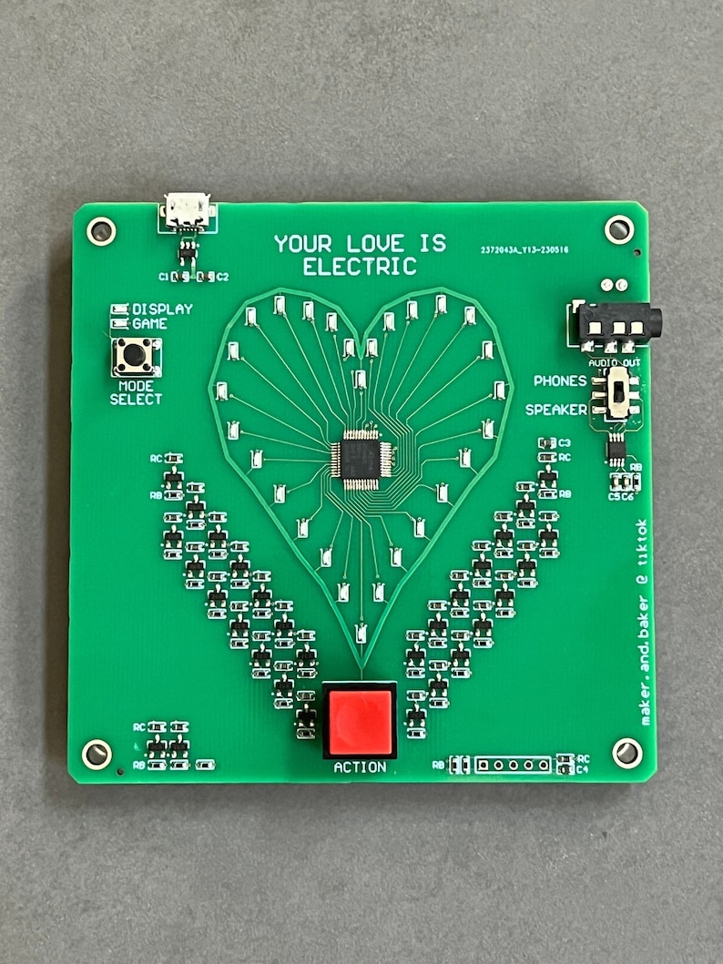 K&ouml;nnte beinhalten: Eine gr&uuml;ne Leiterplatte mit einem herzf&ouml;rmigen Design aus LEDs. Die Platine hat einen roten Knopf mit der Aufschrift "ACTION", einen USB-Anschluss und eine Kopfh&ouml;rerbuchse. Der Text "YOUR LOVE IS ELECTRIC" ist auf der Platine aufgedruckt. Der Text "maker.and.baker @ tiktok" ist auf der rechten Seite der Platine aufgedruckt.
