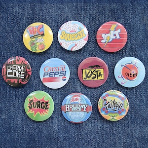 May include: Nine round, colorful, vintage-style buttons with various brand names and logos, including Hi-C, Squeezit, Jolt, Cherry Coke, Crystal Pepsi, Josta, Orbitz, Surge, Holiday Spice, and Frutopia.