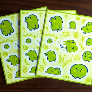 May include: A stack of stickers featuring various green cartoon creatures with blue outlines on a light green background. The stickers have a variety of designs, including duck-like characters, question marks, and wavy lines. The text "WIZEDEN" is also visible.