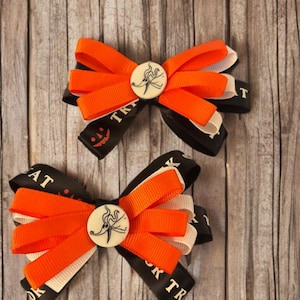 May include: Two hair bows featuring layered orange and cream grosgrain ribbons atop a black ribbon base printed with the text TRICK OR TREAT and a jack-o'-lantern face. Each bow is centered with a circular white button depicting a ghost dog character.