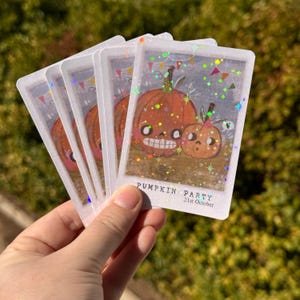 Può includere: Una pila di adesivi rettangolari con finitura olografica a stelle. Ogni adesivo presenta un'illustrazione di due zucche arancioni con volti, festoni colorati e il testo PUMPKIN PARTY 31st October stampato in basso.