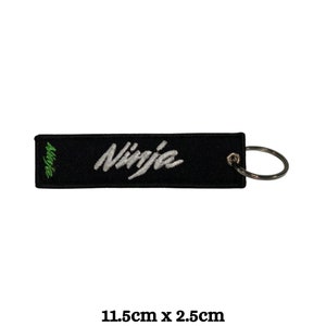 May include: Black fabric keychain with a white "Ninja" logo and a green accent. The keychain measures 1.5 cm by 2.5 cm.