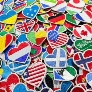 Könnte beinhalten: Eine Sammlung von herzförmigen Stoff-Patches mit Flaggen aus verschiedenen Ländern. Die Patches sind mit weißem Garn genäht und haben eine Vielzahl von Farben, darunter Rot, Weiß, Blau, Grün, Gelb und Schwarz.