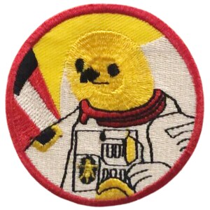 May include: Embroidered patch featuring a yellow cartoon character in a white spacesuit with a red collar. The patch has a red border and a yellow and white background. The character has black eyes and a smiling expression.