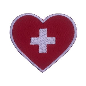 Puede incluir: Parche en forma de corazón rojo con una cruz blanca en el centro. El borde es blanco. El diseño es un símbolo de cuidado y apoyo.