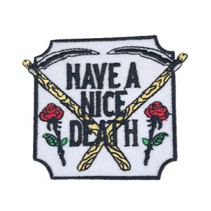 以下が含まれることがあります： 「Have a Nice Death」という黒い文字が書かれた白い刺繍パッチ。パッチには、木製の柄のついた2本の鎌が交差しており、赤いバラが2つ、緑の葉が描かれている。