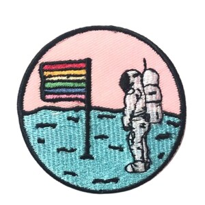 May include: Embroidered patch depicting an astronaut standing on a blue and white surface, planting a rainbow flag against a pink background.