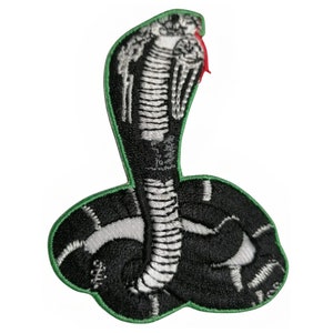 May include: Embroidered patch of a black and white cobra with a green outline. The cobra is in a coiled position with its head raised and its hood expanded. The cobra's eyes are black and its tongue is red.