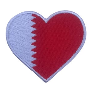 Puede incluir: Un parche rojo en forma de corazón con la bandera de Qatar. La bandera es blanca con una línea roja dentada en el centro.