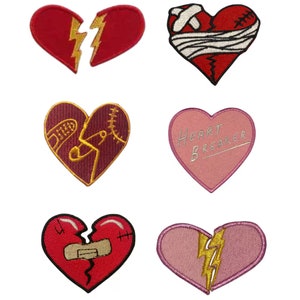May include: Six embroidered iron-on patches in the shape of hearts. The patches feature different designs, including a broken heart with a lightning bolt, a heart with a bandage, a heart with stitches, a heart with the text "Heart Breaker", and a broken heart with a lightning bolt.