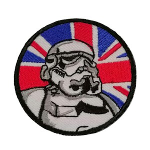 Puede incluir: Un parche blanco bordado con una imagen de dibujos animados de un casco de Stormtrooper delante de una bandera de la Union Jack roja, blanca y azul.