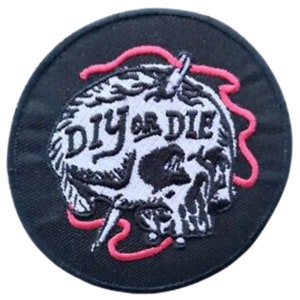 Puede incluir: Parche bordado en blanco y negro con una calavera y el texto "DIY or DIE" en blanco. La calavera está rodeada de un hilo rosa.