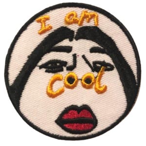 May include: Circular embroidered patch featuring a face design with the text "I am cool". The face has black features, red lips, and the text is in yellow thread on a white background.