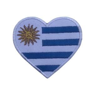 Pode incluir: Um remendo em forma de coração com a bandeira do Uruguai. A bandeira é branca com nove listras horizontais azuis e um sol dourado com um rosto humano no canto superior esquerdo.
