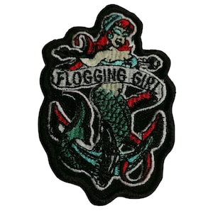 May include: Embroidered patch featuring a mermaid with red hair and a blue cap, holding onto an anchor. The banner above the anchor reads "FLOGGING GIRL". The patch has a black border and is suitable for jackets, bags, or other fabric items.