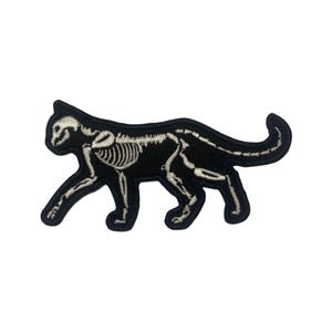 May include: Black embroidered iron-on patch of a cat skeleton in a walking pose. The skeleton is white and the cat is black.