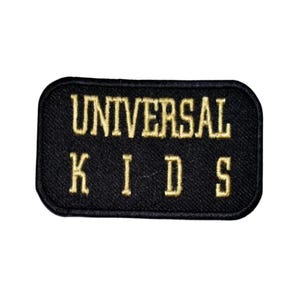 May include: Black rectangular patch with rounded corners. The patch features the words "UNIVERSAL KIDS" embroidered in gold thread. The patch is likely designed to be sewn or ironed onto clothing or accessories.