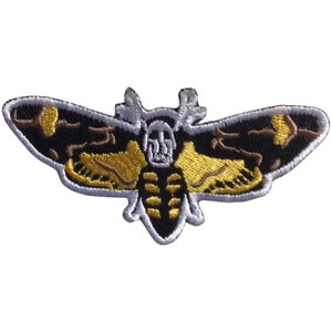 Puede incluir: Un parche bordado para planchar con un diseño de esfinge de la muerte de color negro, marrón y amarillo.