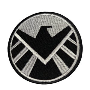May include: A black and white embroidered patch featuring the S.H.I.E.L.D. logo. The logo is a circle with a white eagle inside and a white star in the centre. The eagle has its wings spread out and its head is turned to the right. The star is in the centre of the eagle's chest.