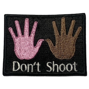 May include: Black embroidered patch with two hands, one pink and one brown, raised in the air with the text "Don't Shoot" in white.