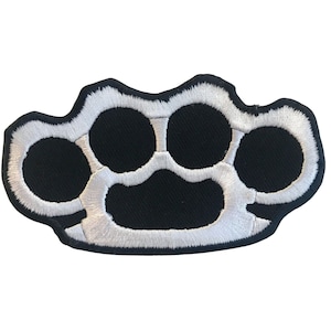May include: A white embroidered patch in the shape of a brass knuckle. The patch is on a black background.