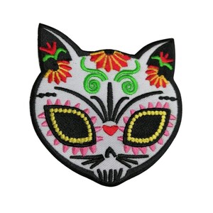 Könnte beinhalten: Weißer gestickter Katzenkopf-Patch mit schwarzen, rosa, gelben, grünen und roten Details. Die Augen der Katze sind schwarz mit gelben Umrandungen und das Gesicht hat ein schädelartiges Design.
