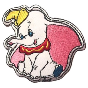 Puede incluir: Un parche bordado de Dumbo el elefante de Disney. Dumbo está sentado con las orejas levantadas y lleva un traje de circo rojo y amarillo. El parche tiene un fondo rosa y un contorno gris.