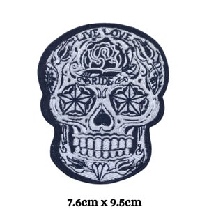 May include: Black and white embroidered iron-on patch featuring a sugar skull with a star-shaped eye design. The skull has a floral design around it and the text "Live Love Ride" above it. The patch measures 7.6 cm by 9.5 cm.
